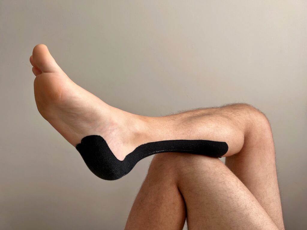 Close-up of a person's foot with black kinesiology tape applied to the heel and arch, used for treating plantar fasciitis, heel pain, or supporting the foot during sports injury recovery